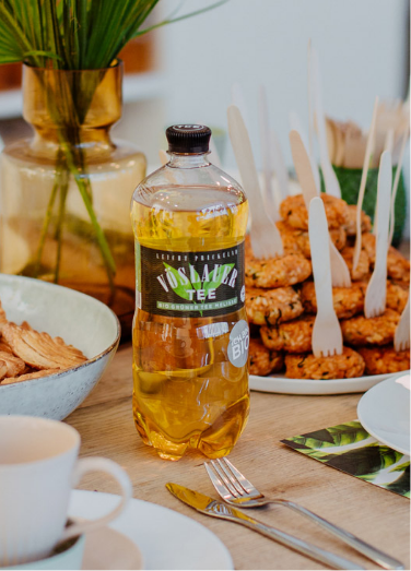 Ein gedeckter Tisch mit Snacks im Hintergrund, im Fokus ist eine Vöslauer Tee Flasche
