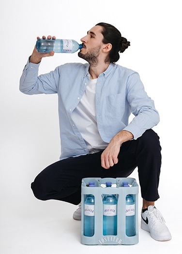 Mann hockt vor einer Kist Vöslauer Mineralwasser und trinkt aus einer Flasche